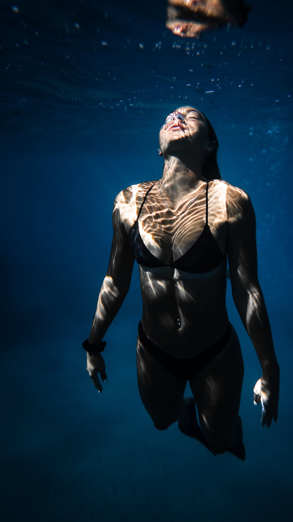 Underwater shot of fit young female in black bikini swimming in dark blue seawater with eyes closed